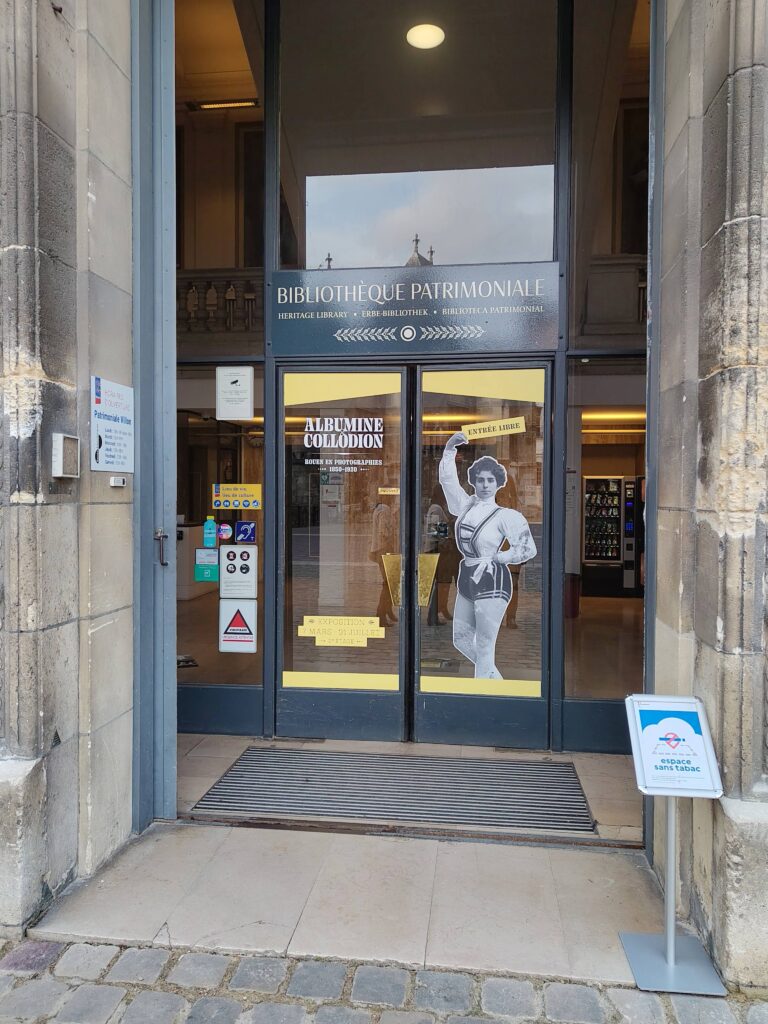 Photographie de l'entrée de la Bibliothèque patrimoniale. Sur la porte, il y a le nom de l'exposition ainsi que la photographie d'une danseuse du Théâtre des arts de Rouen du 19ème siècle